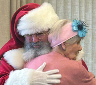 A person dressed as Santa Claus warmly hugs a senior wearing a pink sweater and a pink headband with a blue flower in front of beige curtains.

Description generated by AI