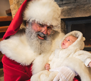 A person dressed as Santa Claus with glasses and a white beard is smiling and holding a newborn wrapped in a fluffy white blanket near a stone fireplace.

Description generated by AI