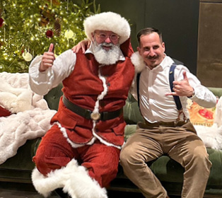 A person dressed as Santa Claus with glasses and a white beard smiles while holding a child wearing a festive sweater in front of a decorated Christmas tree with lights.

Description generated by AI