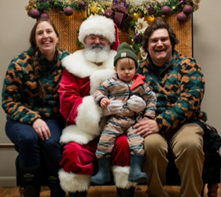 A child wearing a striped outfit and green beanie sits on the lap of a person dressed as Santa Claus while another person in a patterned fleece jacket sits beside them smiling.

Description generated by AI